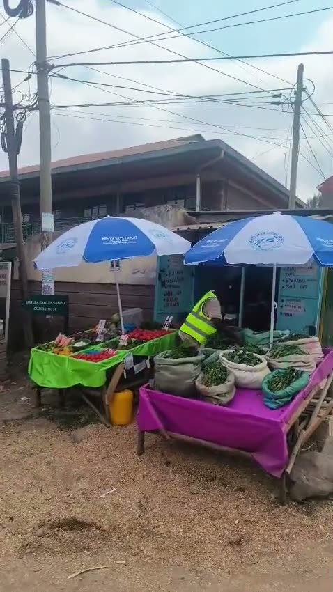 A vendor in a hi-vis vest serving customers from a Kenya Skys Credit branded produce stall